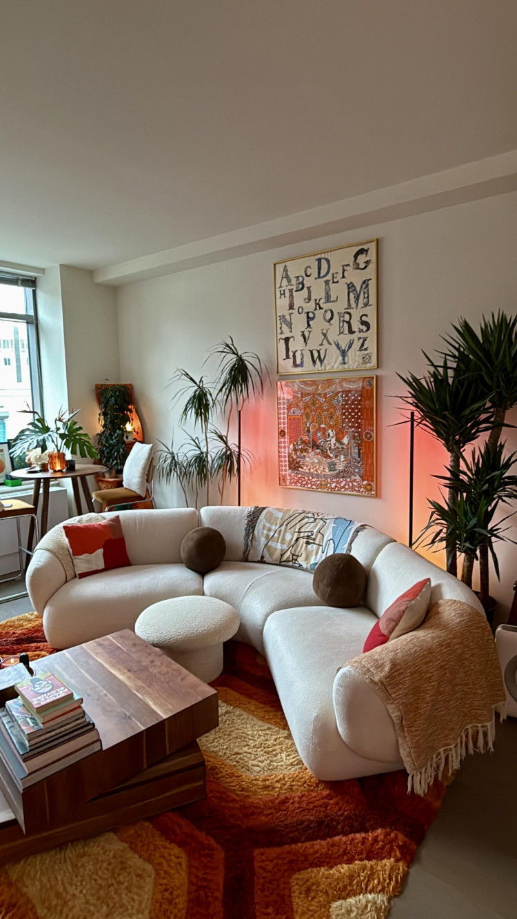 Mid-century modern living room with a curved white sofa, textured orange and brown rug, wooden coffee table, green plants, and warm ambient lighting.