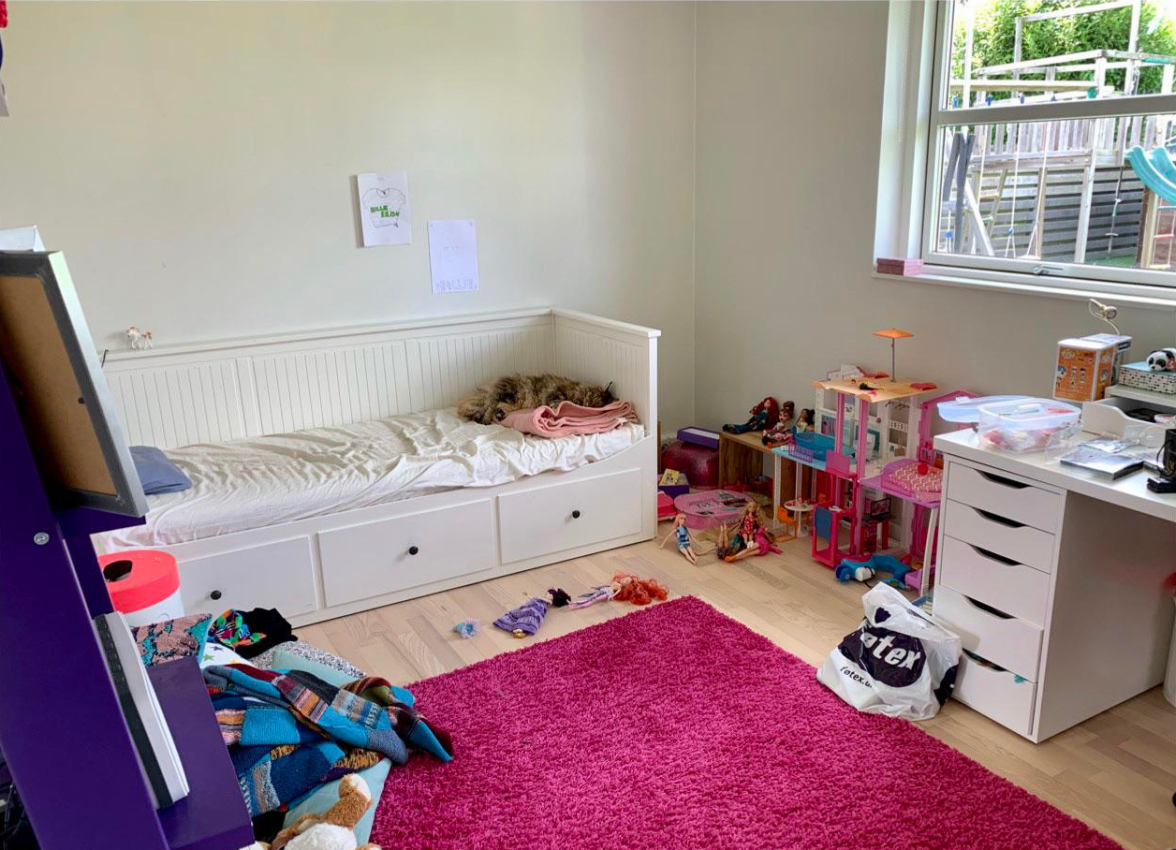 Kids bedroom with white daybed, pink rug, toy clutter, desk area, and dollhouse beside a bright window