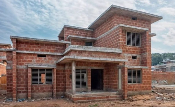 An unfinished two-story brick house with concrete lintels and pillars, rectangular windows with metal bars, under cloudy sky, construction site atmosphere.