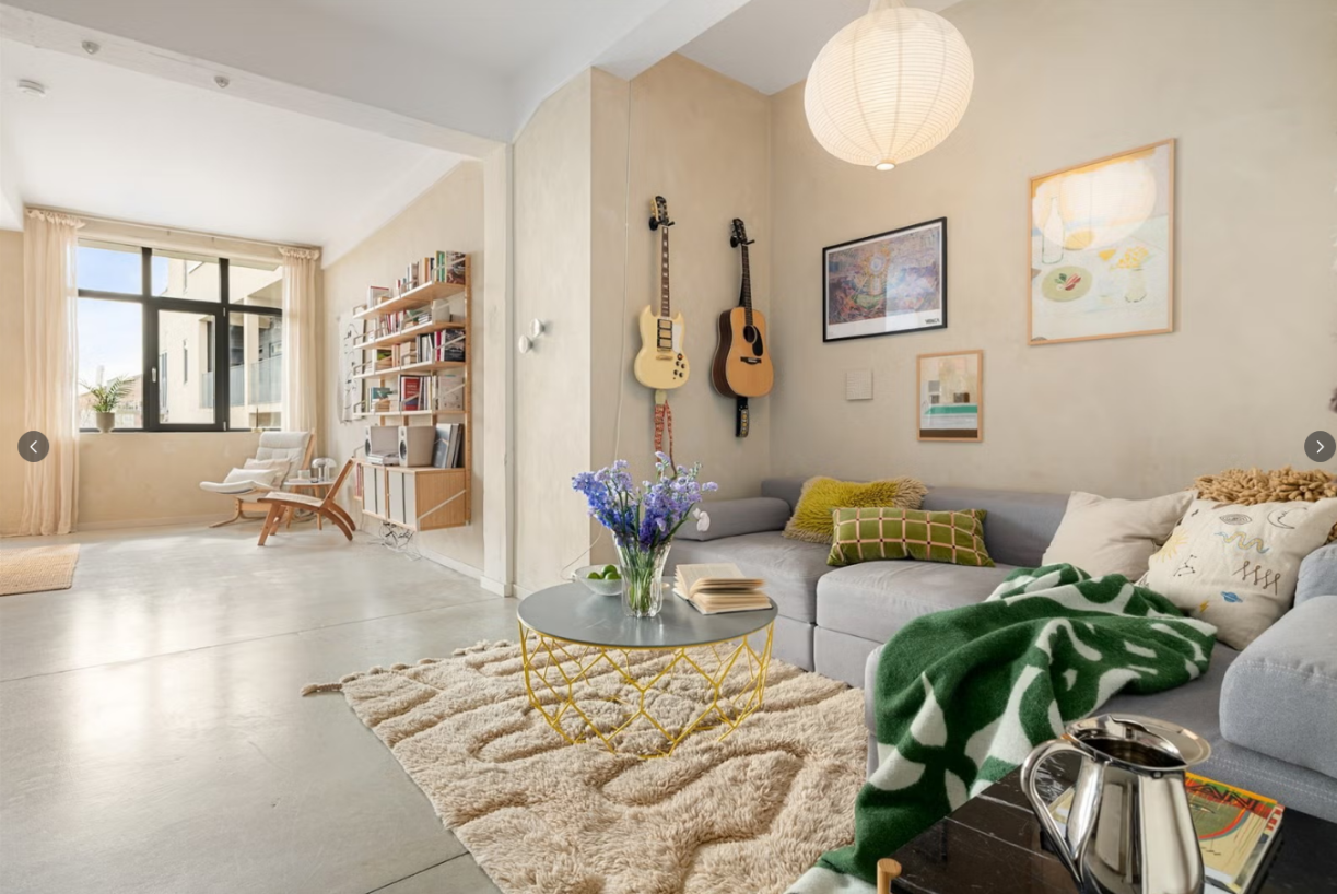 Bright contemporary living room with beige walls, gray sectional sofa, colorful pillows, hanging guitars, round coffee table, shaggy rug, and large window.