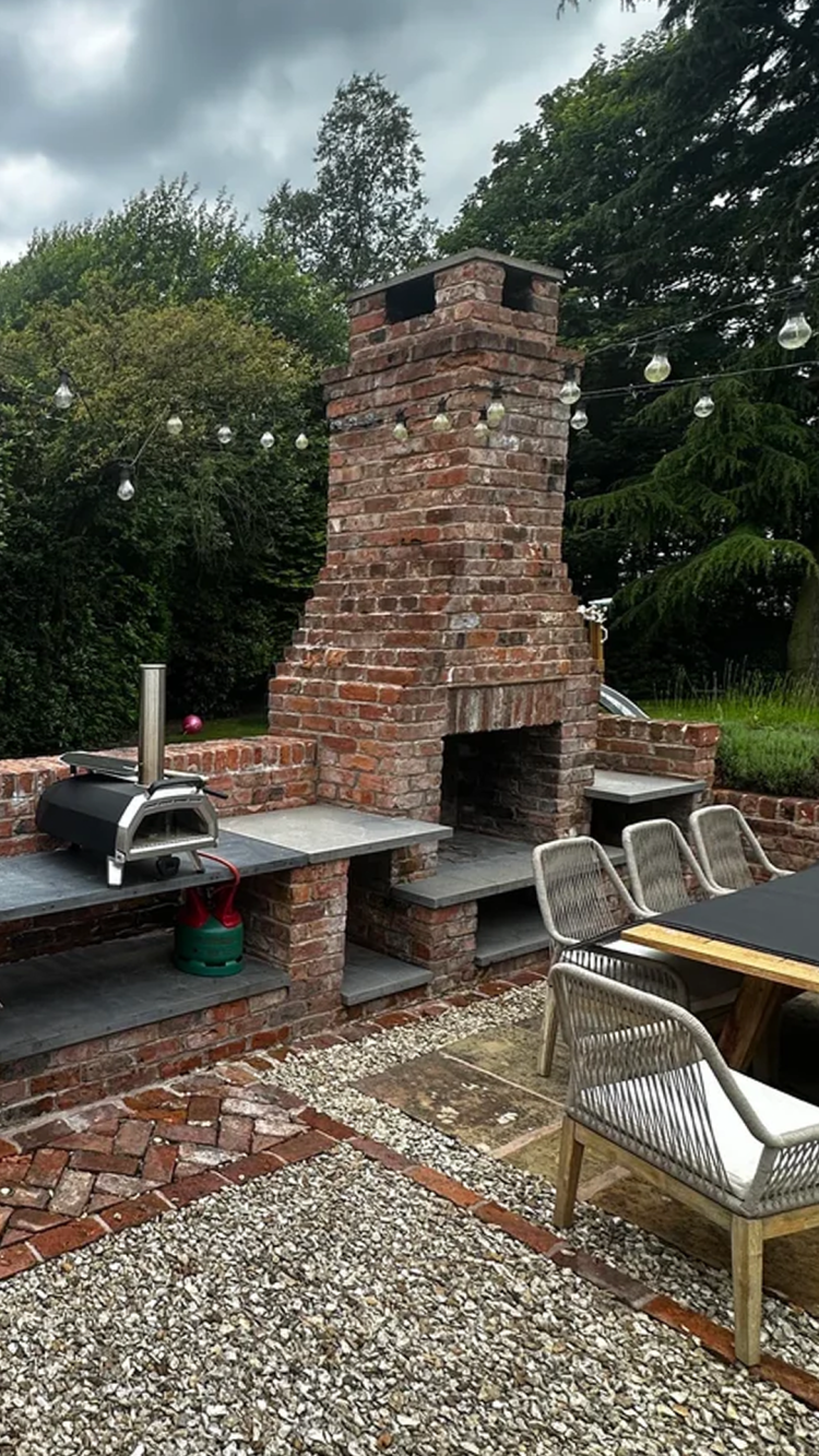 An outdoor rustic patio with large brick fireplace, string lights overhead, stone and brick flooring, woven chairs, and a modern pizza oven, surrounded by greenery.