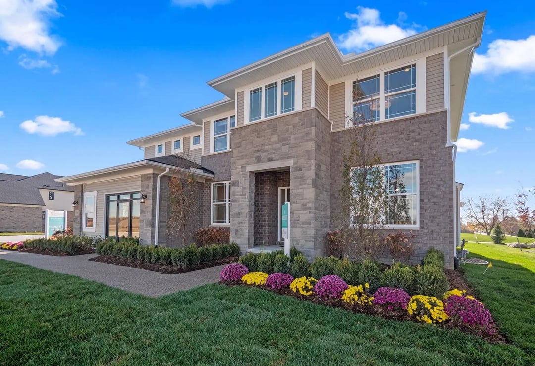 Two story suburban house with gray brick exterior, white trim, landscaped flower beds, and large front windows