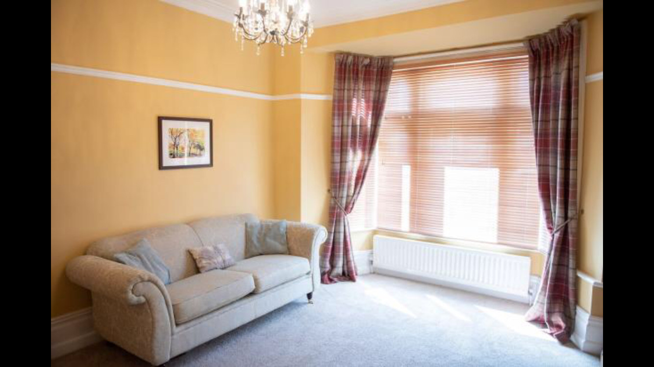 A Living room with traditional style, featuring beige walls, a light gray rolled-arm sofa, plaid red curtains, wooden blinds.