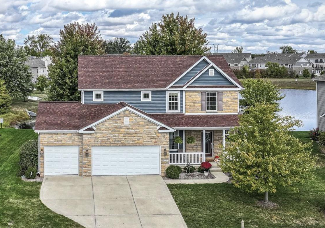 Two-story suburbaBlue suburban farmhouse exterior with stone facade and attached garagen house with blue siding and beige stone facade, three-car garage, front porch with plants, surrounded by green lawn and trees, under cloudy sky.