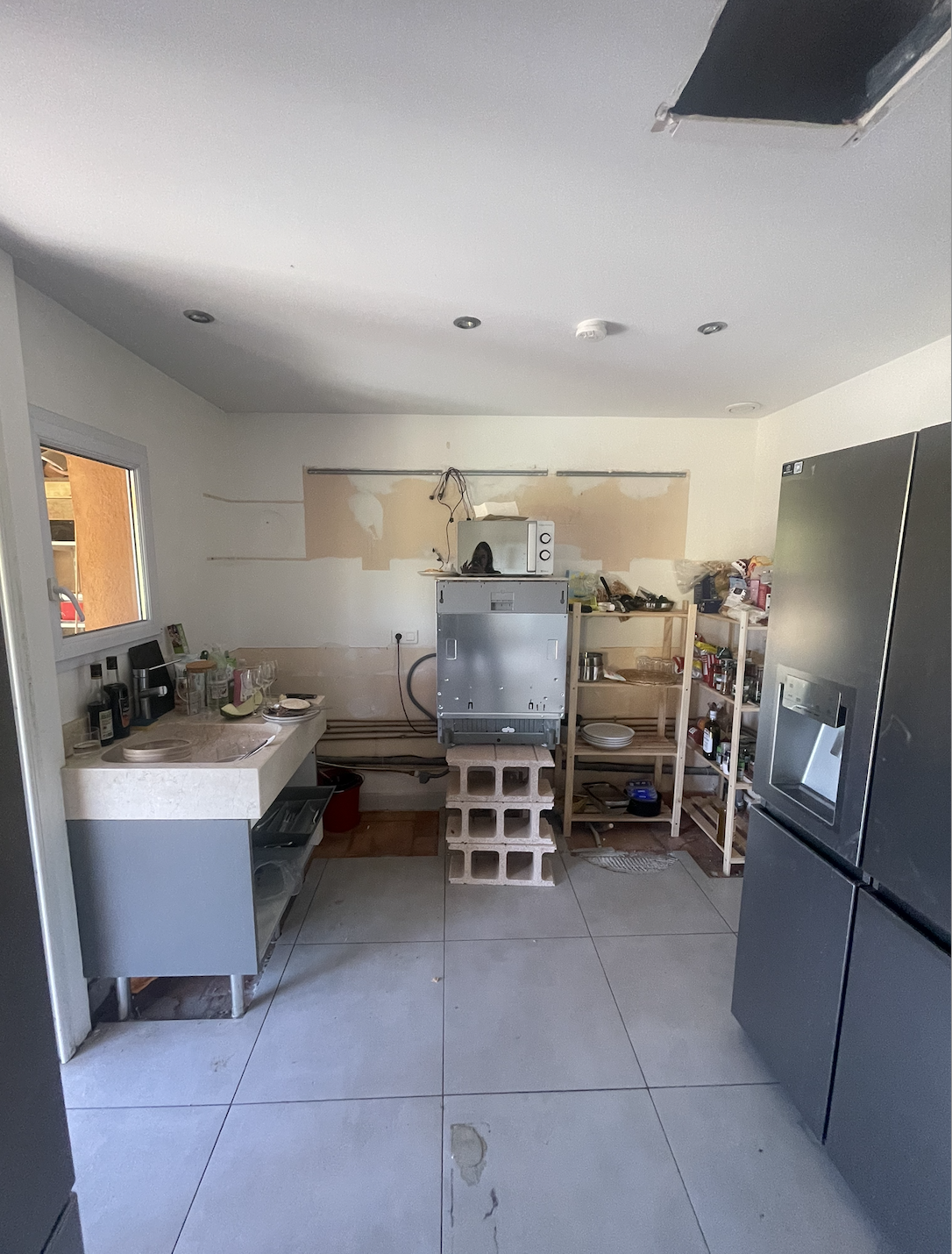 A cluttered kitchen with unfinished walls, gray tiled floor, minimalist gray cabinetry, exposed appliances on cinder blocks, wooden shelving, and a cluttered, utilitarian atmosphere.