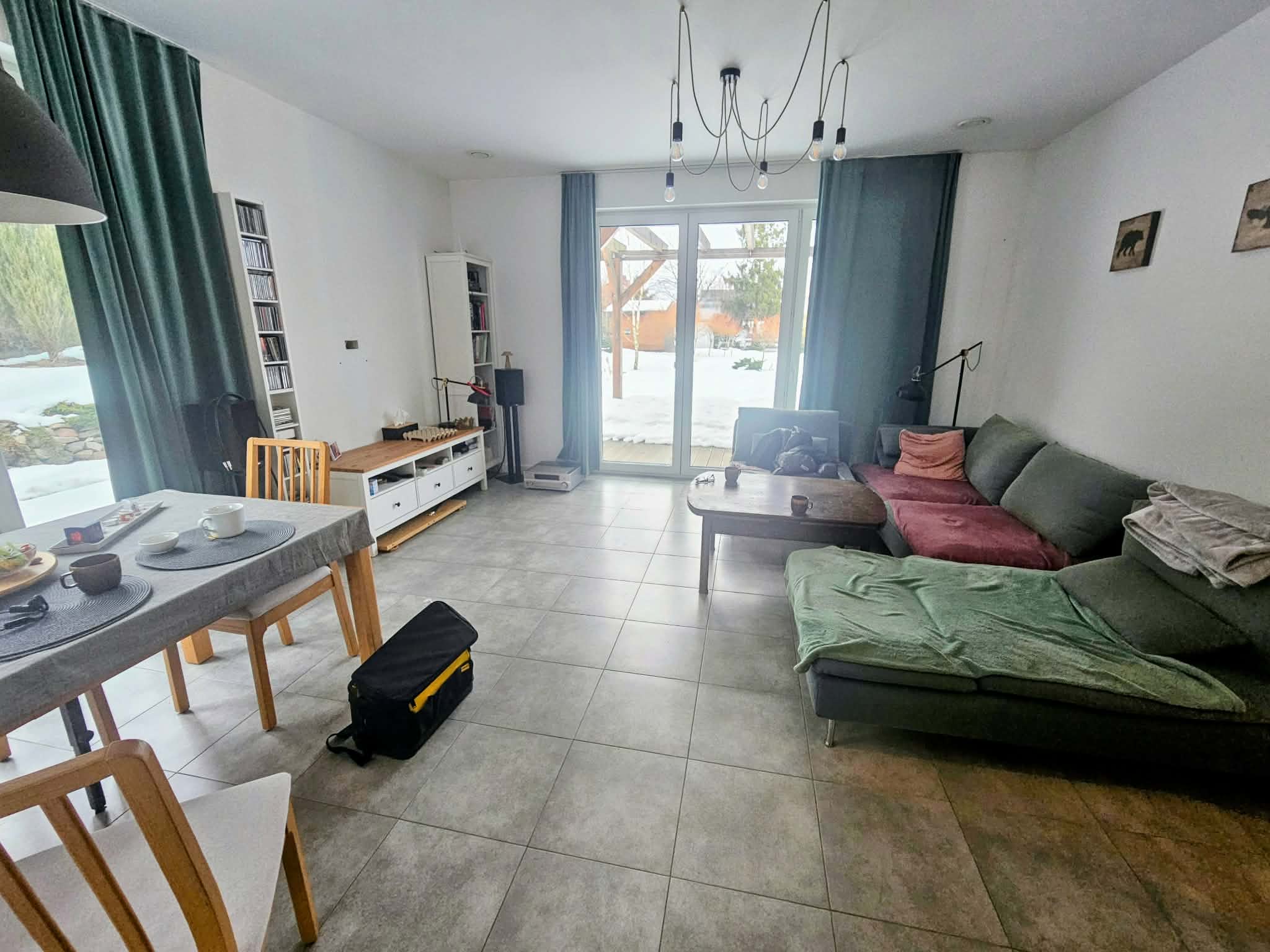 Dated living room with modern minimalist style, featuring green and pink sofa, wooden dining table, large windows with teal curtains, and neutral tiled floor.