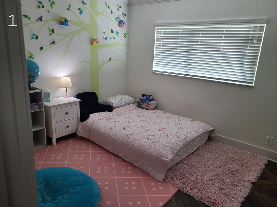 Kids bedroom with tree wall mural, pink rugs, floor mattress, and white nightstand beside a large window