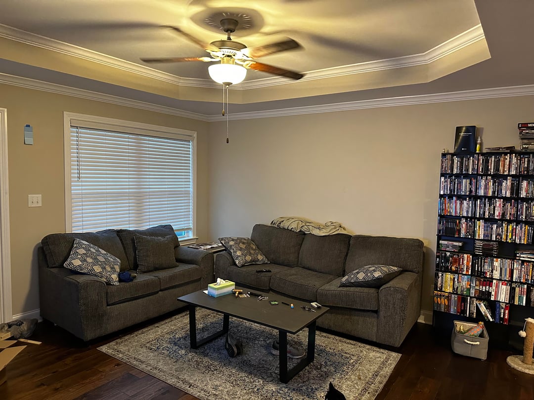 Traditional living room with gray fabric sofas, patterned pillows, dark wood floors, a black coffee table, large window with blinds, and a ceiling fan.