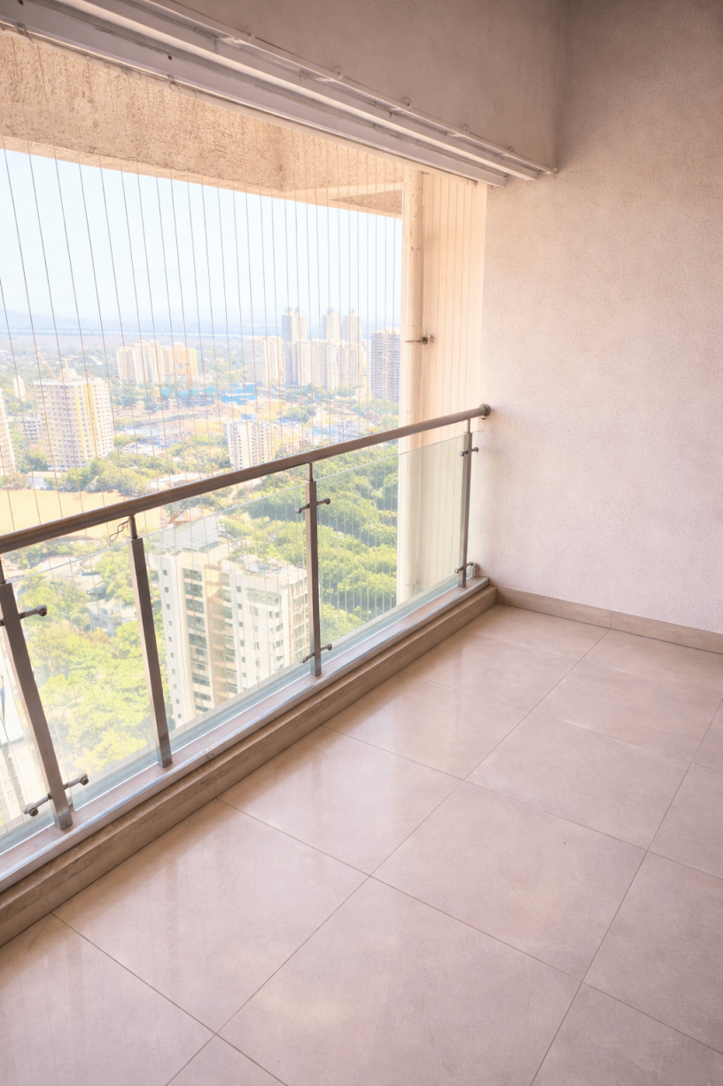 Empty modern balcony with beige tiled floor, glass and metal railing, textured light walls, overlooking a cityscape with high-rise buildings under bright daylight.