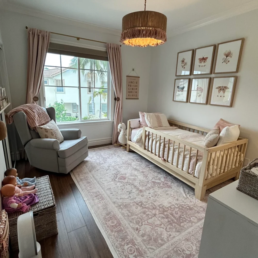 Kids bedroom with natural wood daybed, soft pink and beige tones, butterfly wall art, plush armchair, large window, and patterned area rug.