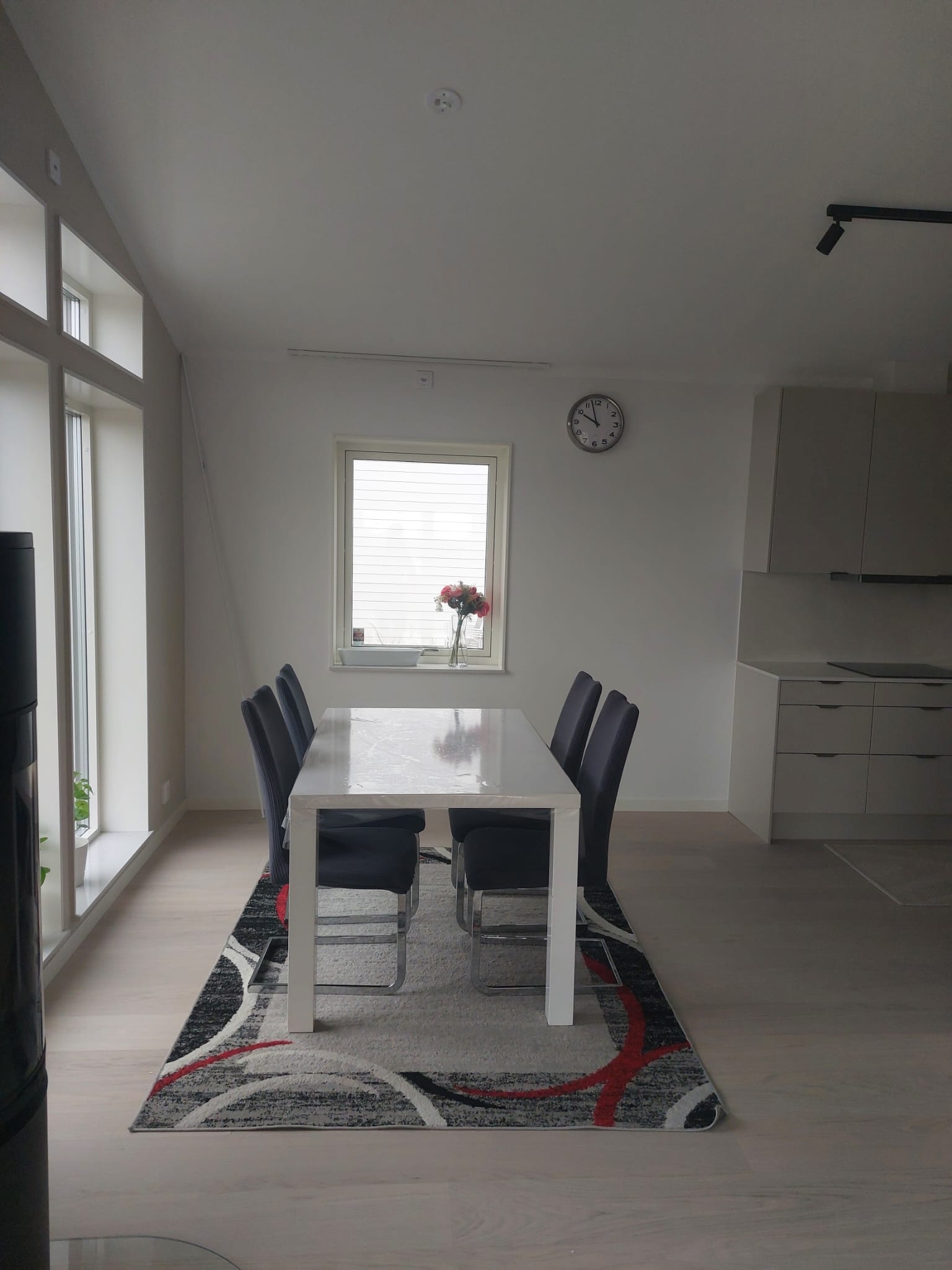 A dark kitchen dining area with white walls, black dining chairs, a white table on a patterned rug, small window with flowers, and neutral cabinetry.