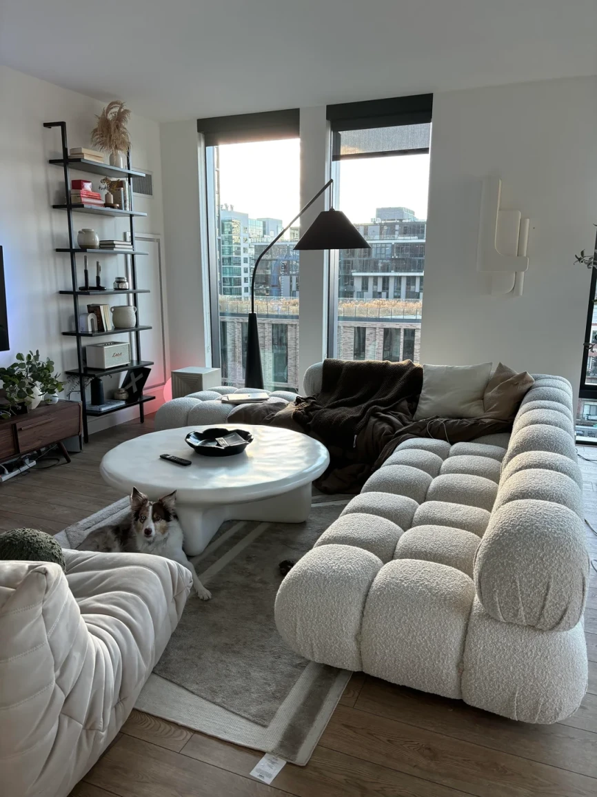 Modern living room with tufted white boucle sofa, round white coffee table, black floor lamp, tall shelving unit, large windows, and cozy neutral tones.