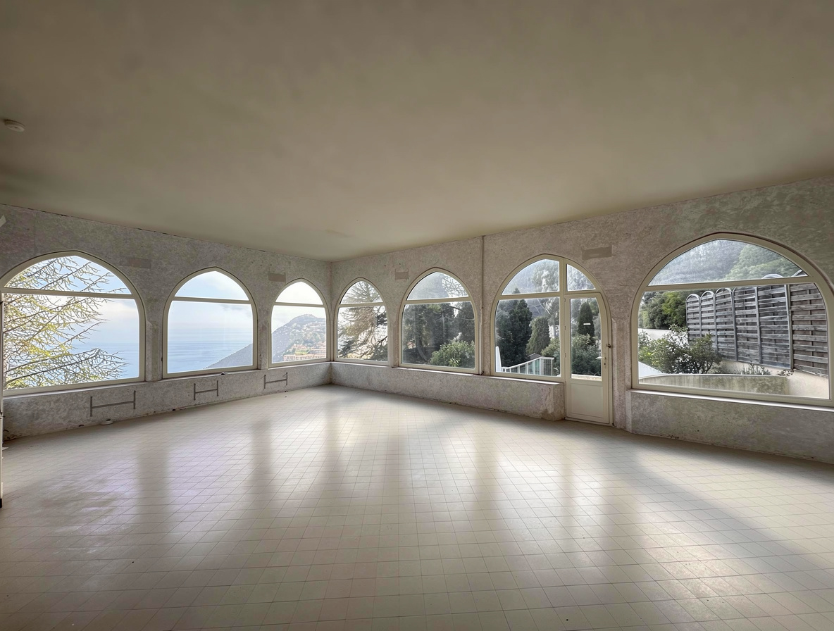 An empty open-concept room with tiled flooring, textured walls, and multiple large arched windows overlooking a waterfront and hillside view.