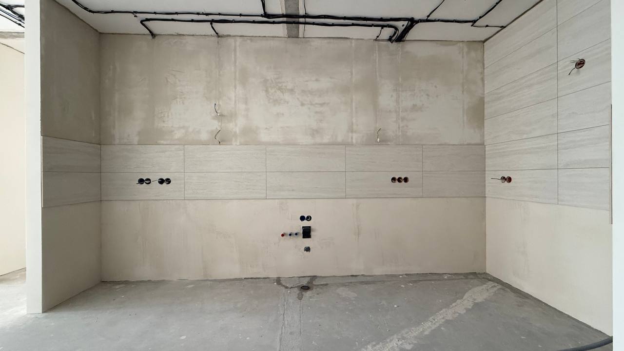 An empty unfinished kitchen space with bare concrete floor, gray tiled walls, exposed wiring, and plumbing fixtures, showcasing a raw, industrial, minimalistic atmosphere.