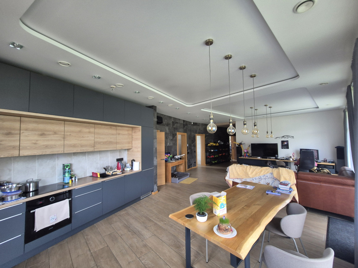Spacious modern kitchen and living area with dark gray and wood cabinetry, natural wooden dining table, pendant lights, and neutral tones.
