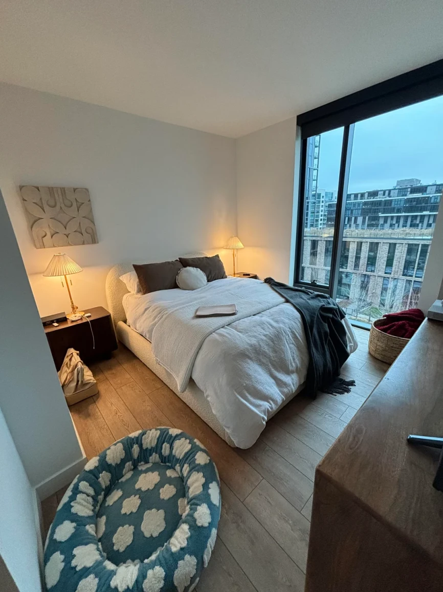 Minimalist modern bedroom with neutral tones, white bedding, dark accent pillows, wooden floor, soft lighting from bedside lamps, large window with city view.
