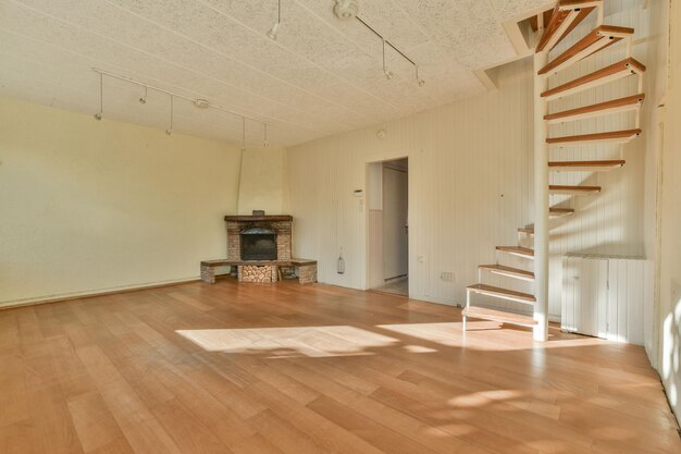 Spacious living room with light wood flooring, cream walls, rustic stone fireplace, and white spiral staircase, featuring natural sunlight and minimalist style.