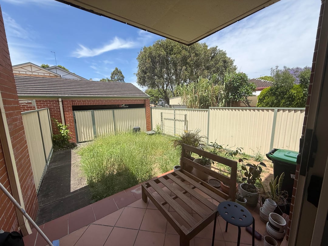Overgrown courtyard backyard with patchy grass, tiled patio, and side pathway