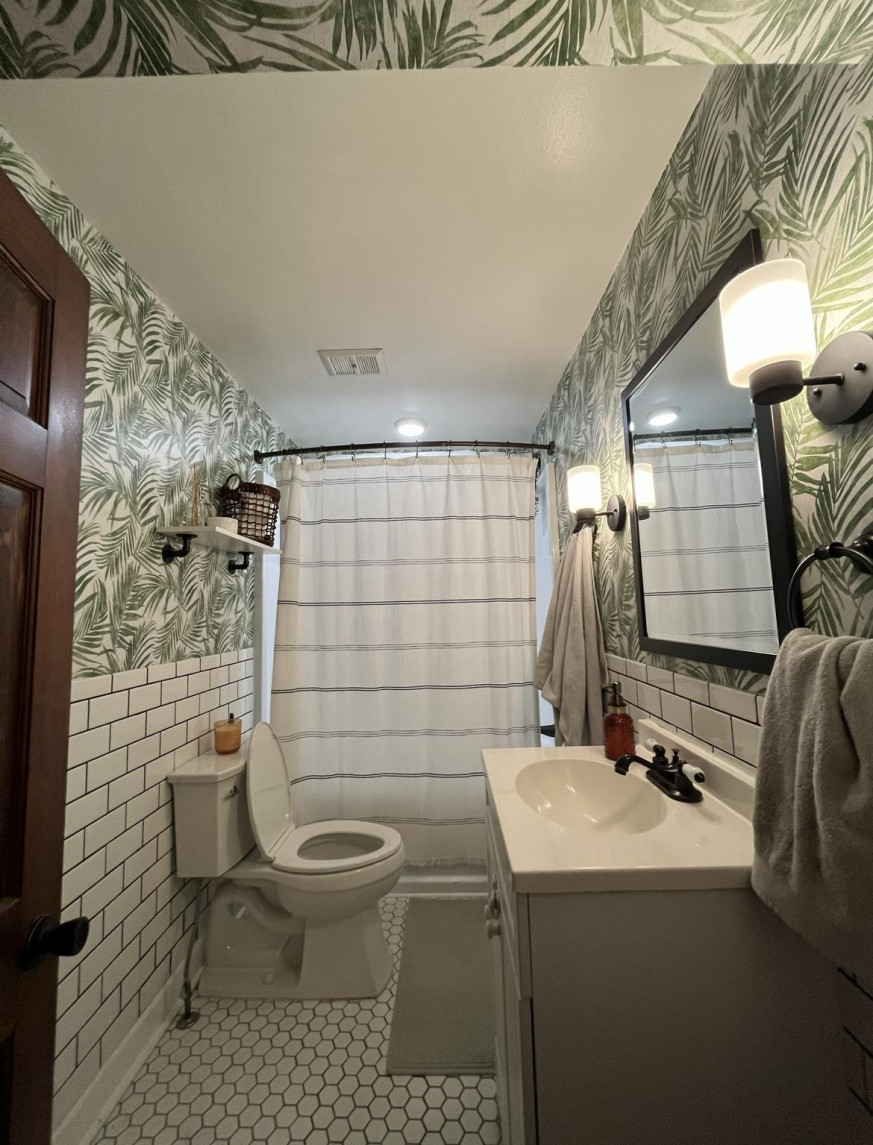 Bathroom with tropical leaf wallpaper, white subway tiles, black framed mirror, and striped shower curtain