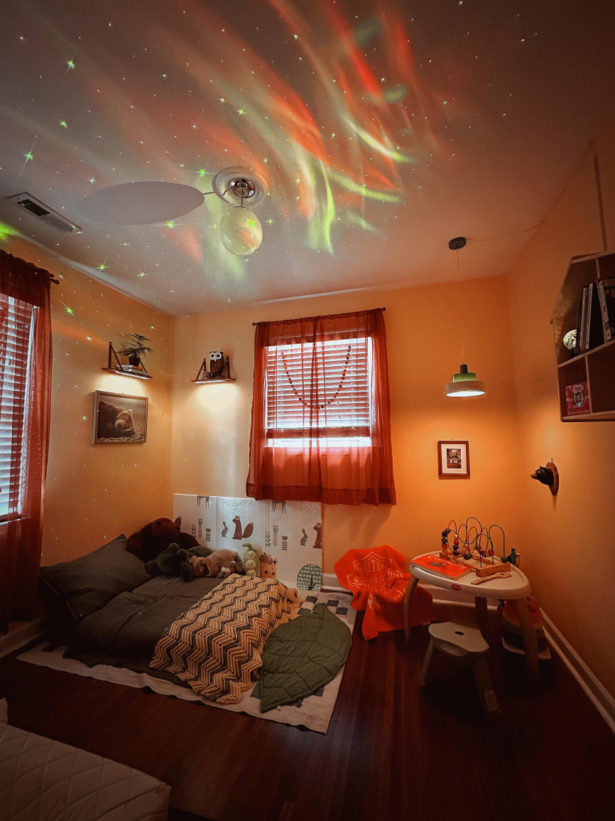 Toddler bedroom with orange walls, floor mattress, play table, dark curtains, and galaxy ceiling light effects
