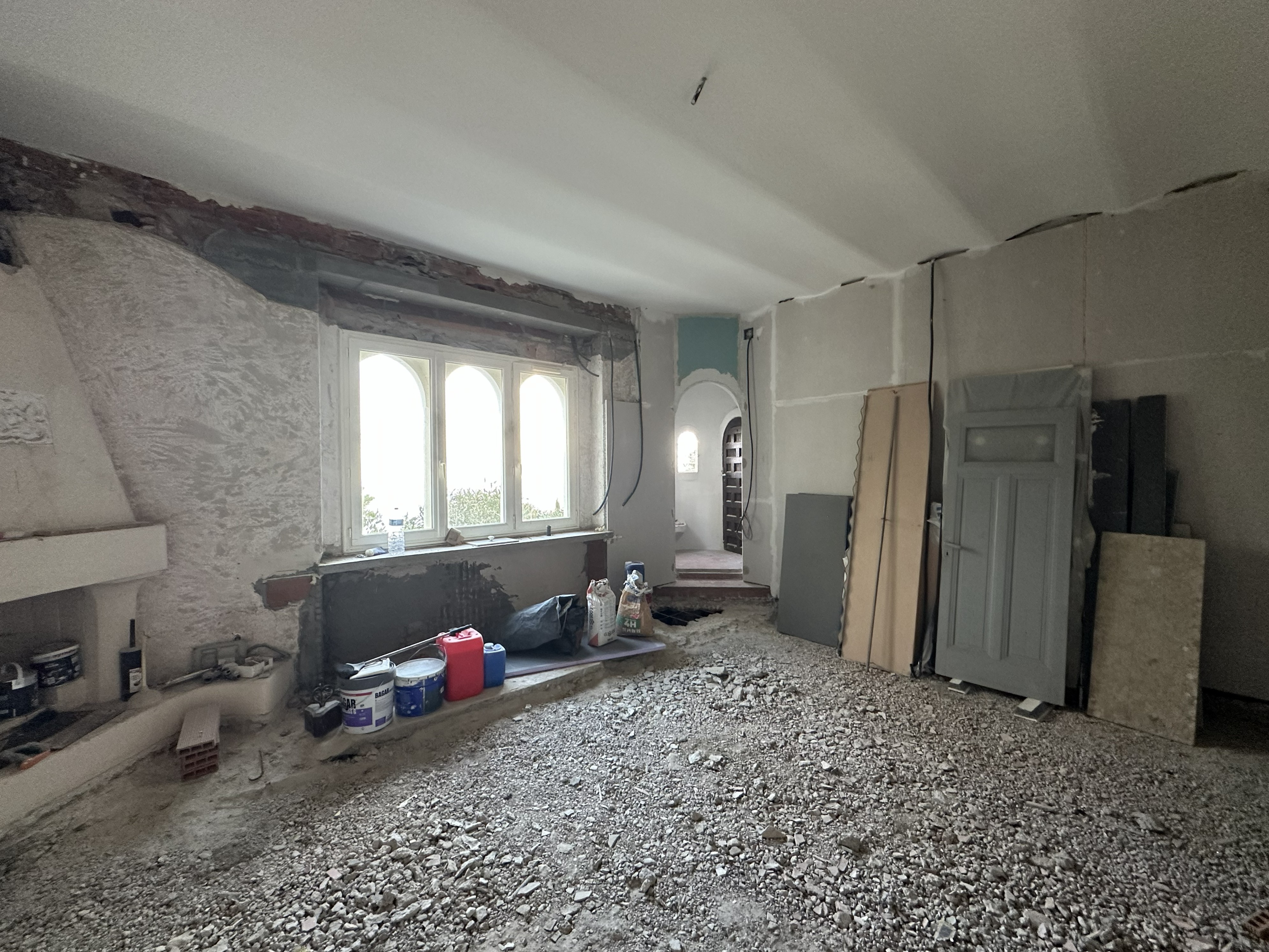 Bedroom under renovation with exposed stone walls, arched window, rough gravel floor, construction materials, and unfinished plaster, creating a raw, industrial atmosphere.