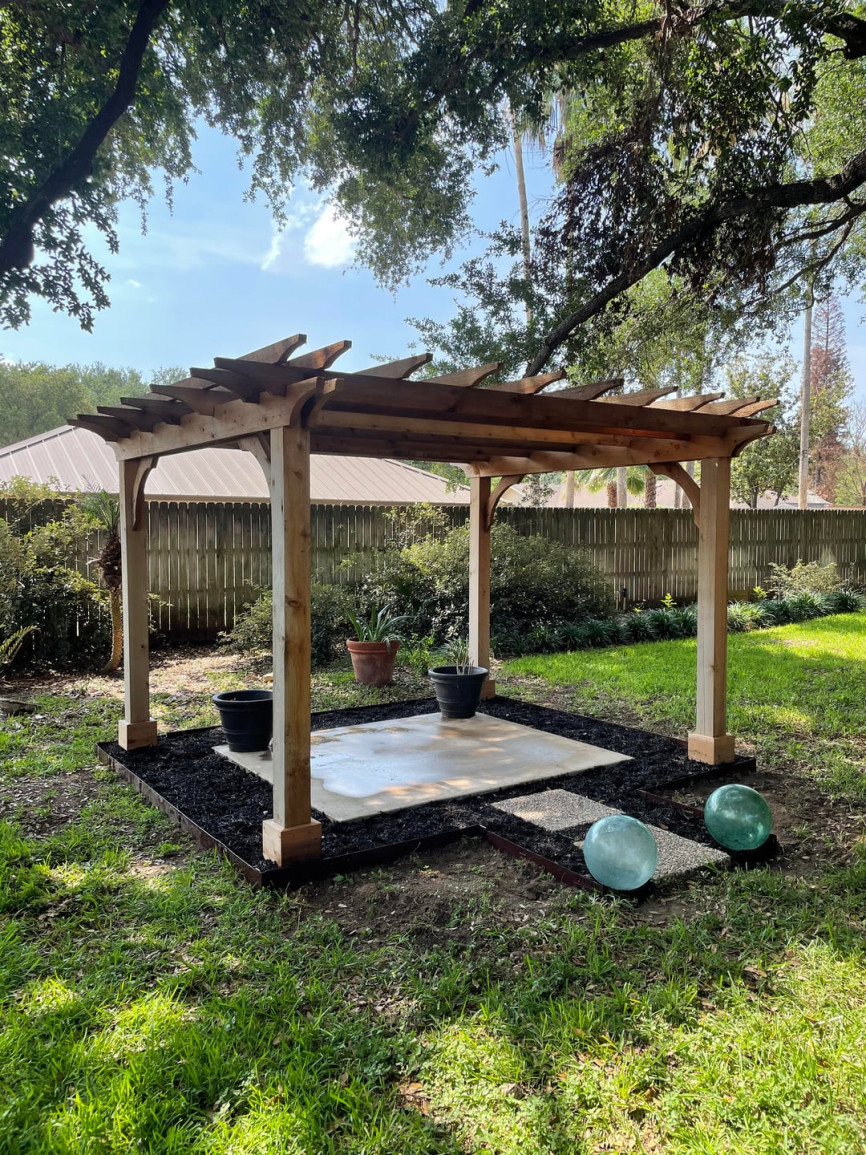 Outdoor backyard pergola with natural wood beams over a concrete patio, surrounded by green grass, potted plants, and shaded by large trees.
