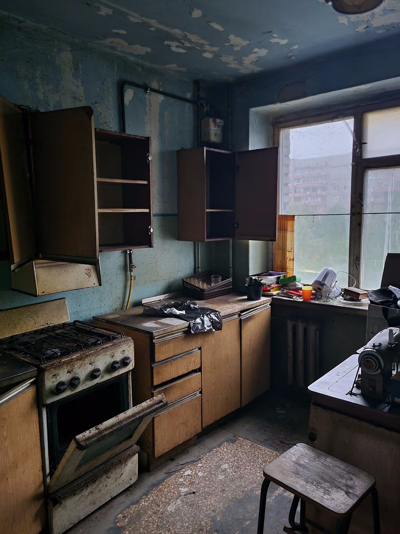 A dim, worn-out kitchen with peeling blue walls, old wooden cabinets with open doors, rusty stove, cluttered windowsill, and a vintage sewing machine, evoking a neglected, dated atmosphere.
