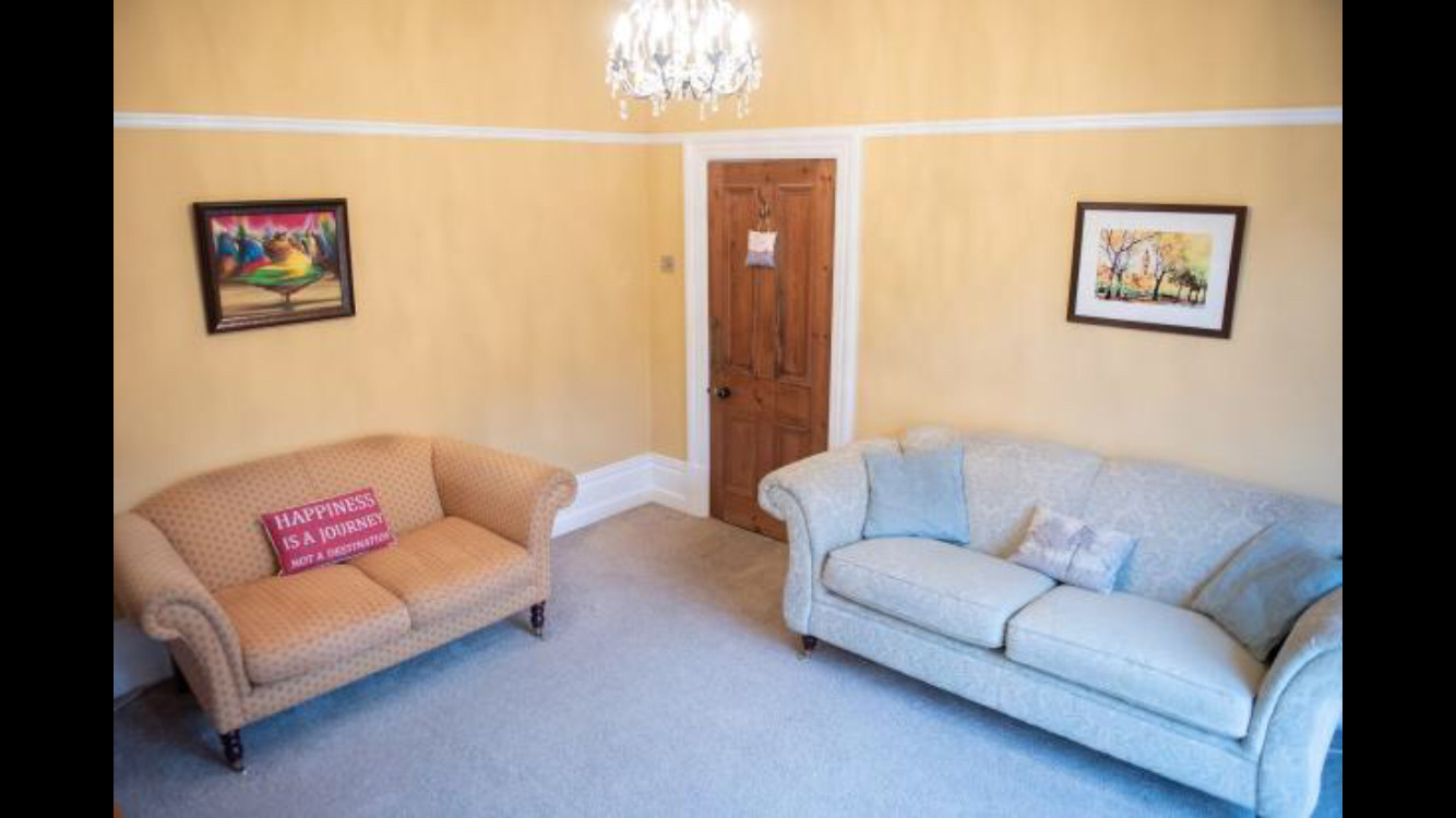 Living room with traditional style, featuring beige and light blue patterned sofas, warm yellow walls, framed landscape paintings, wooden door, and soft lighting.