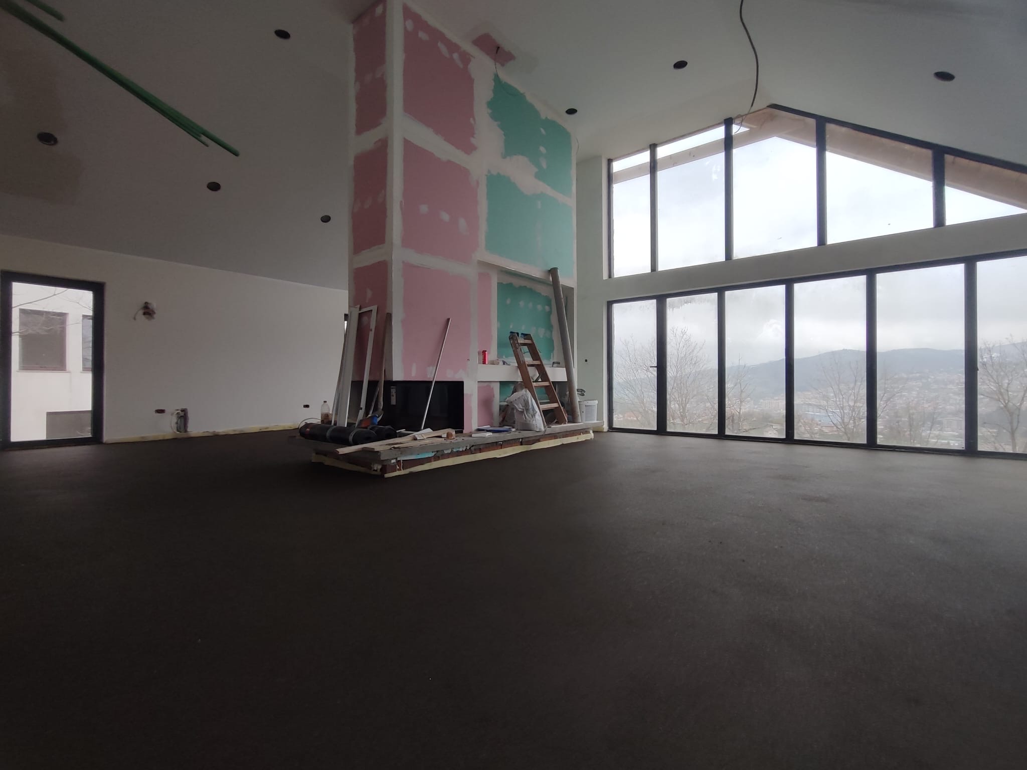 Spacious modern living room under construction with large floor-to-ceiling windows, high ceilings, exposed drywall in pink and teal, and minimal natural light.