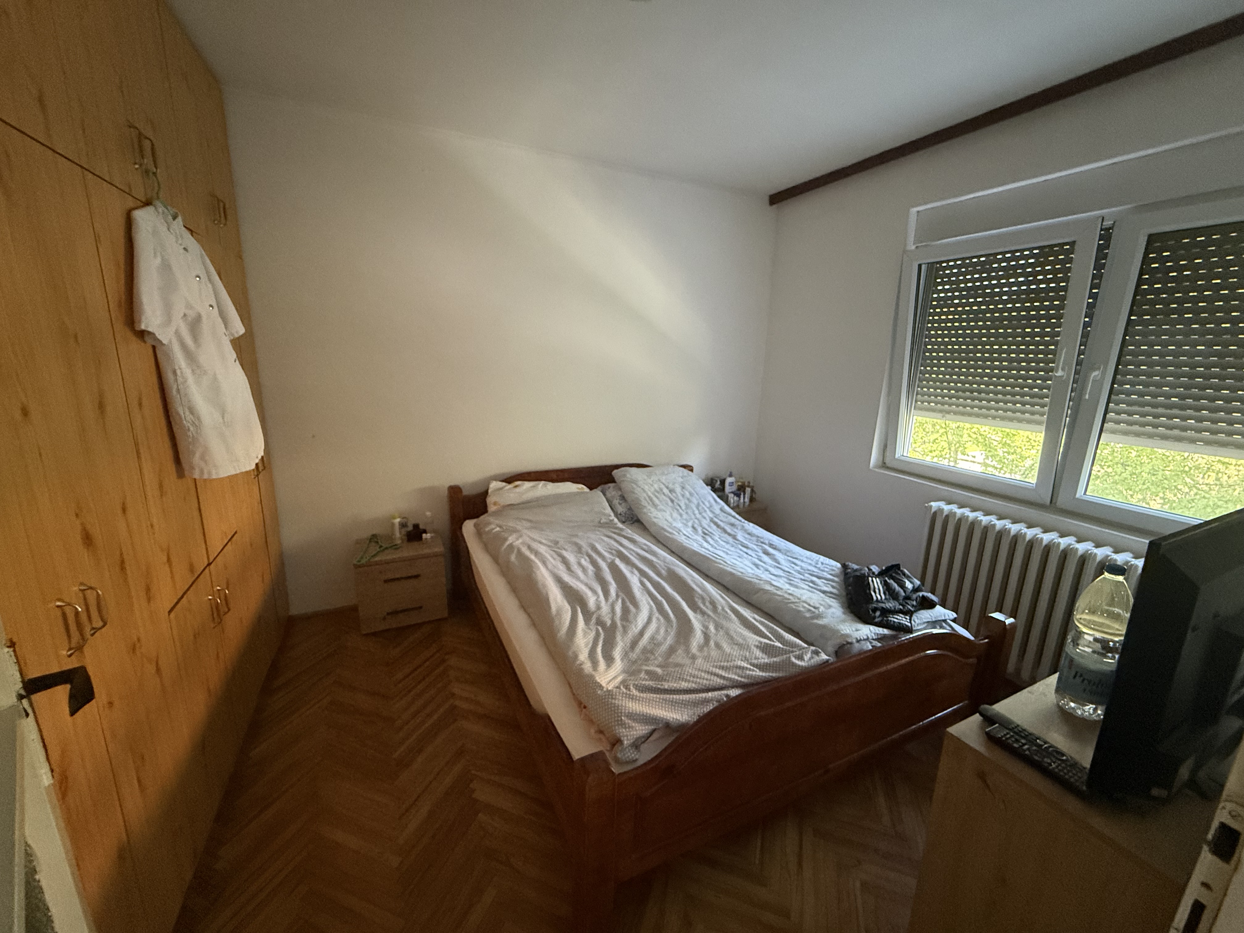 Small, dated bedroom with wooden parquet floor, large wooden wardrobe, unmade bed with white bedding, closed window blinds, and minimal natural light.
