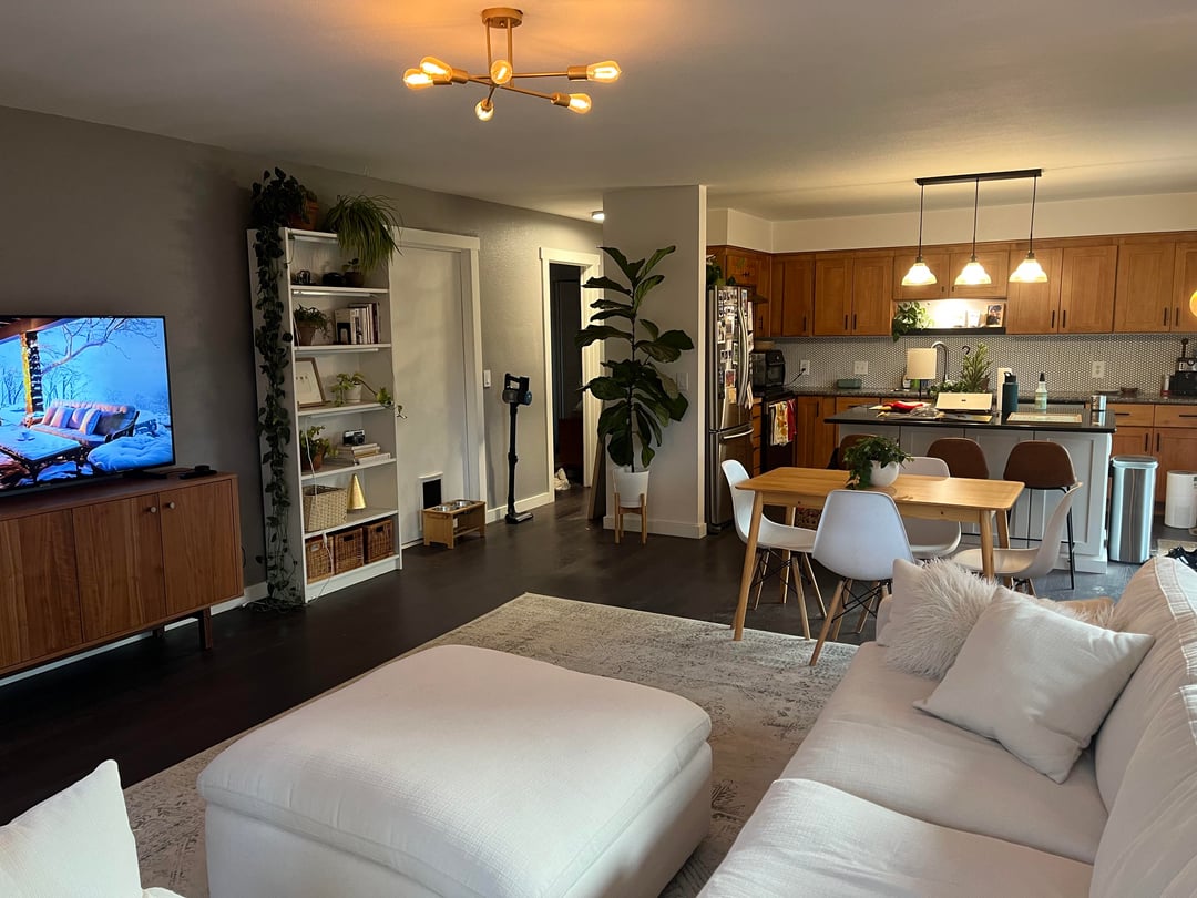 Open-concept living room with sectional sofa, rectangular dining table, dark flooring, and wood kitchen cabinetry.