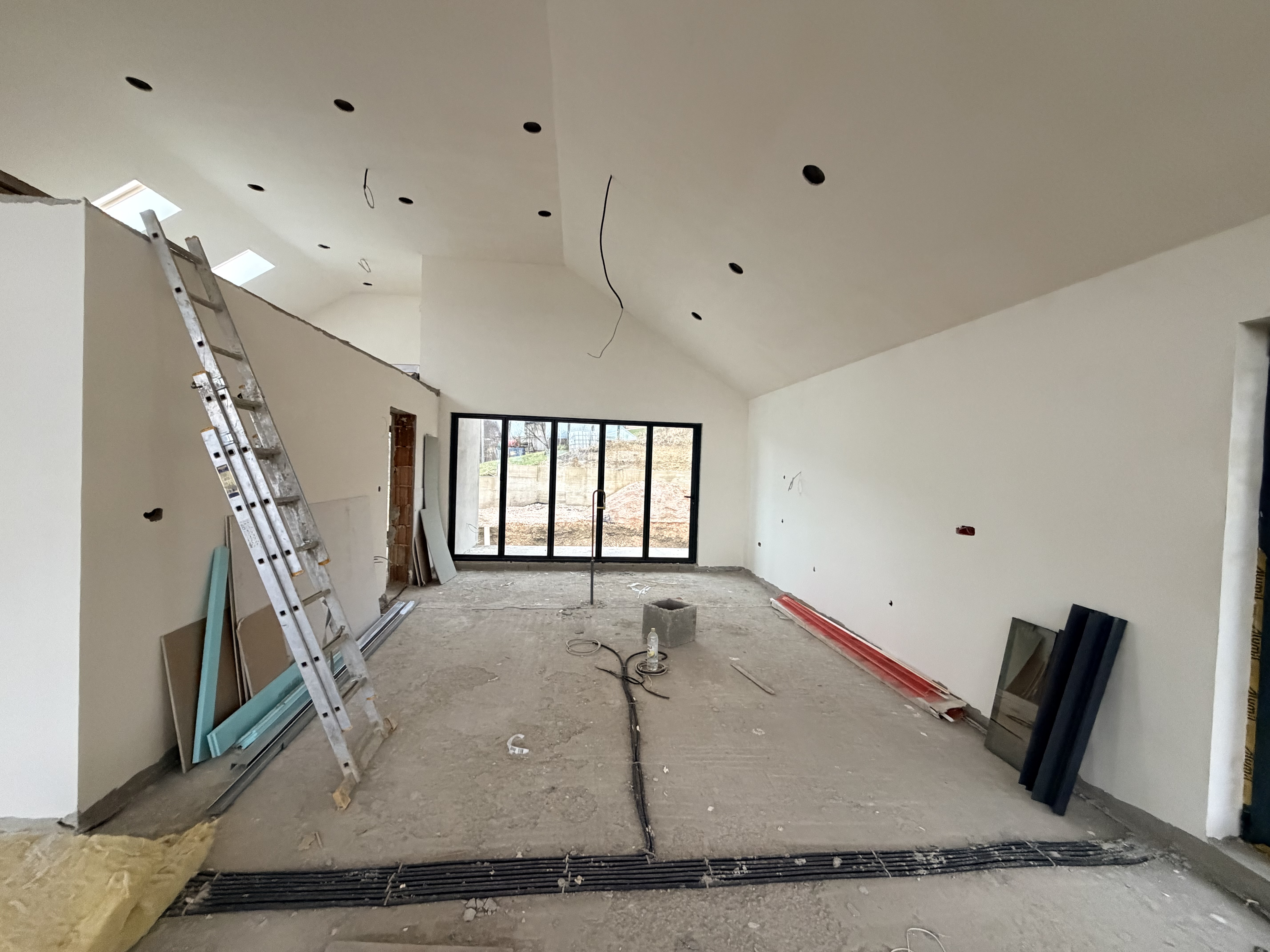 Spacious kitchen under construction with high vaulted ceilings, white walls, exposed wiring, ladder, large black-framed sliding doors, and natural light.