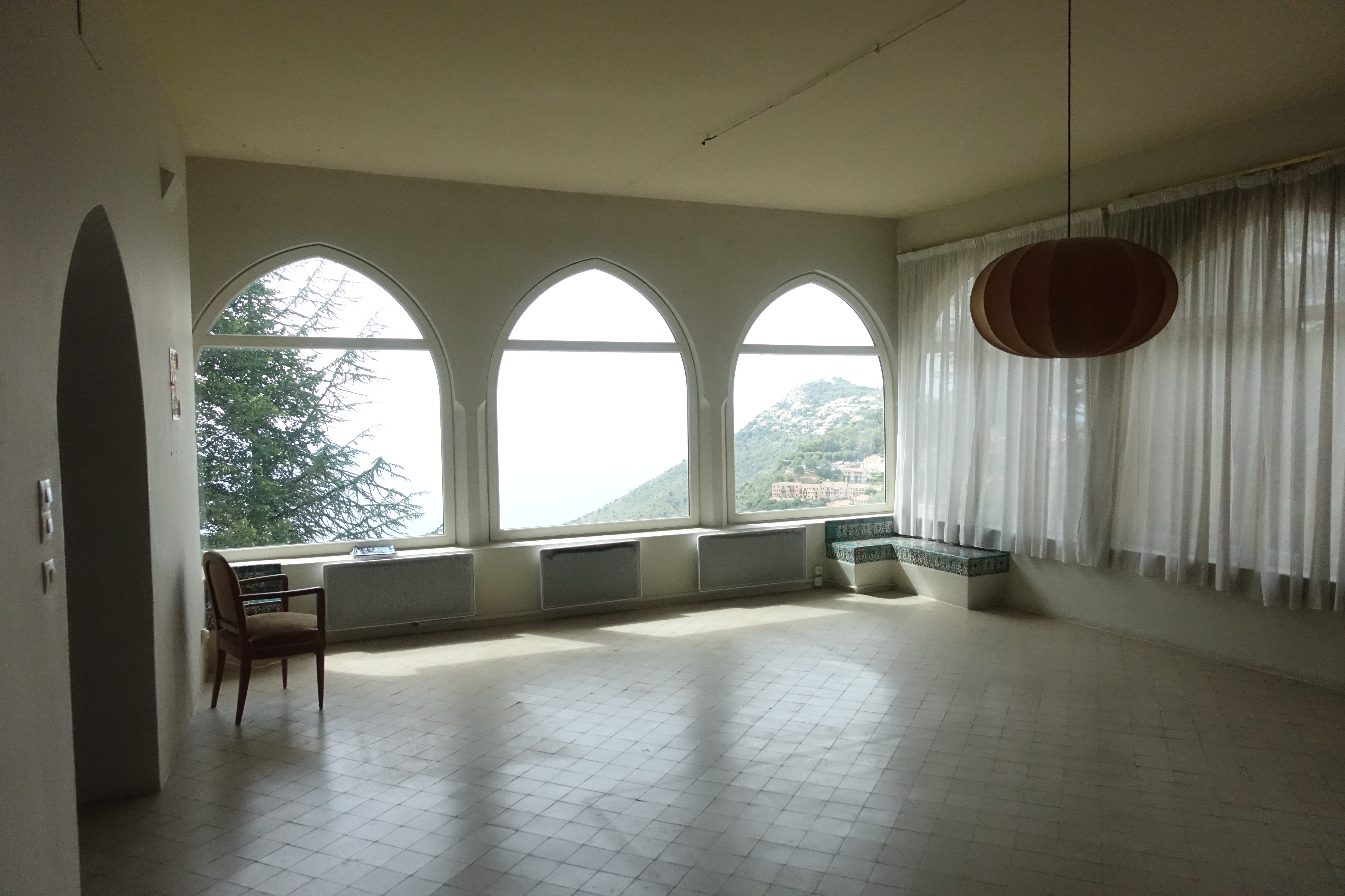 Empty living room with Mediterranean style, featuring large arched windows, tiled floor, built-in corner bench with mosaic accents, sheer white curtains, and a wooden pendant light.