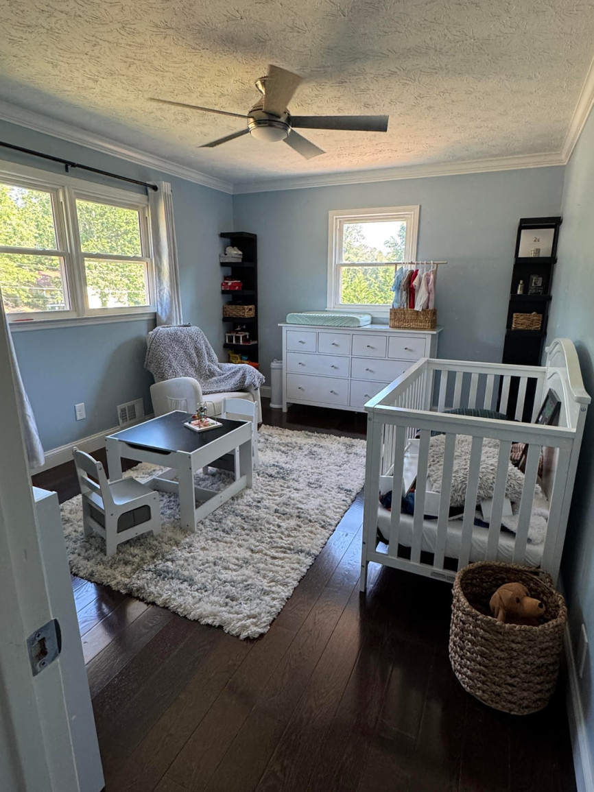 Toddler bedroom with crib, white dresser, shag rug, small activity table, and blue walls with natural light.