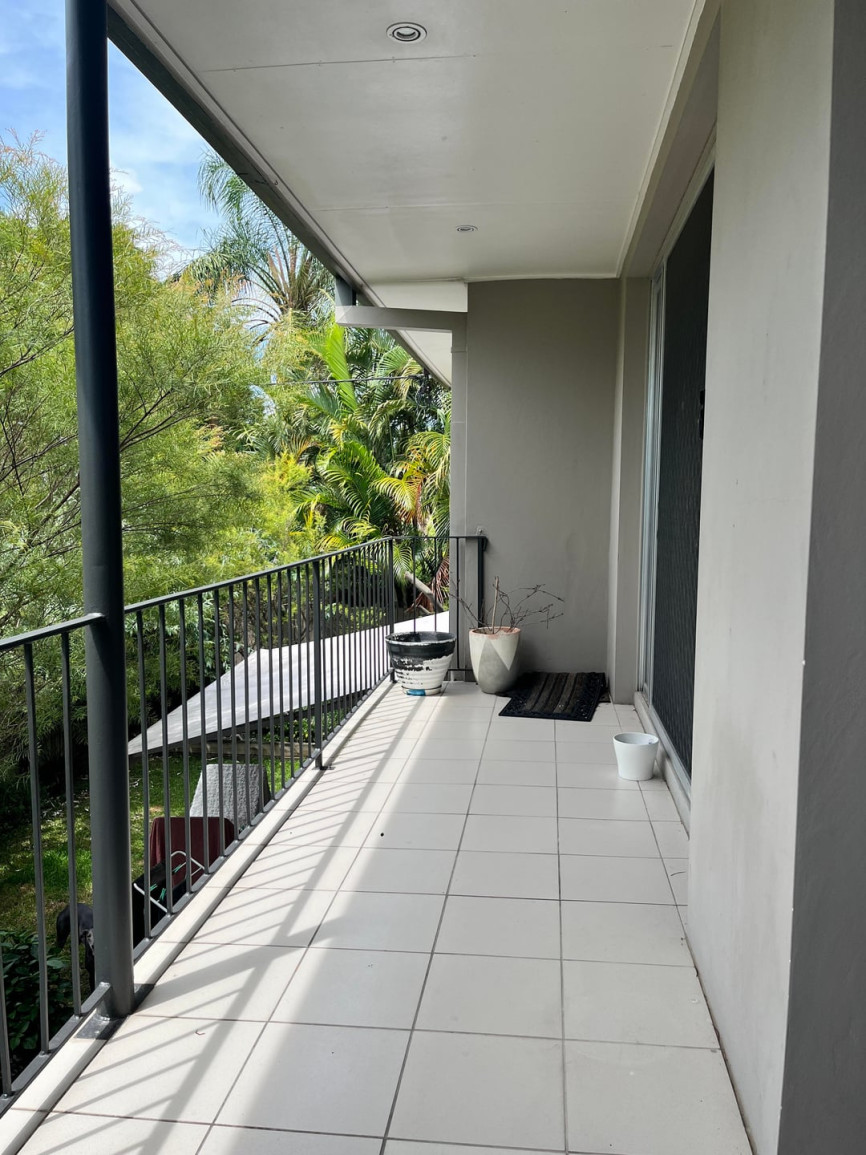 Covered apartment balcony with tiled floor, black railing, and surrounding tropical trees