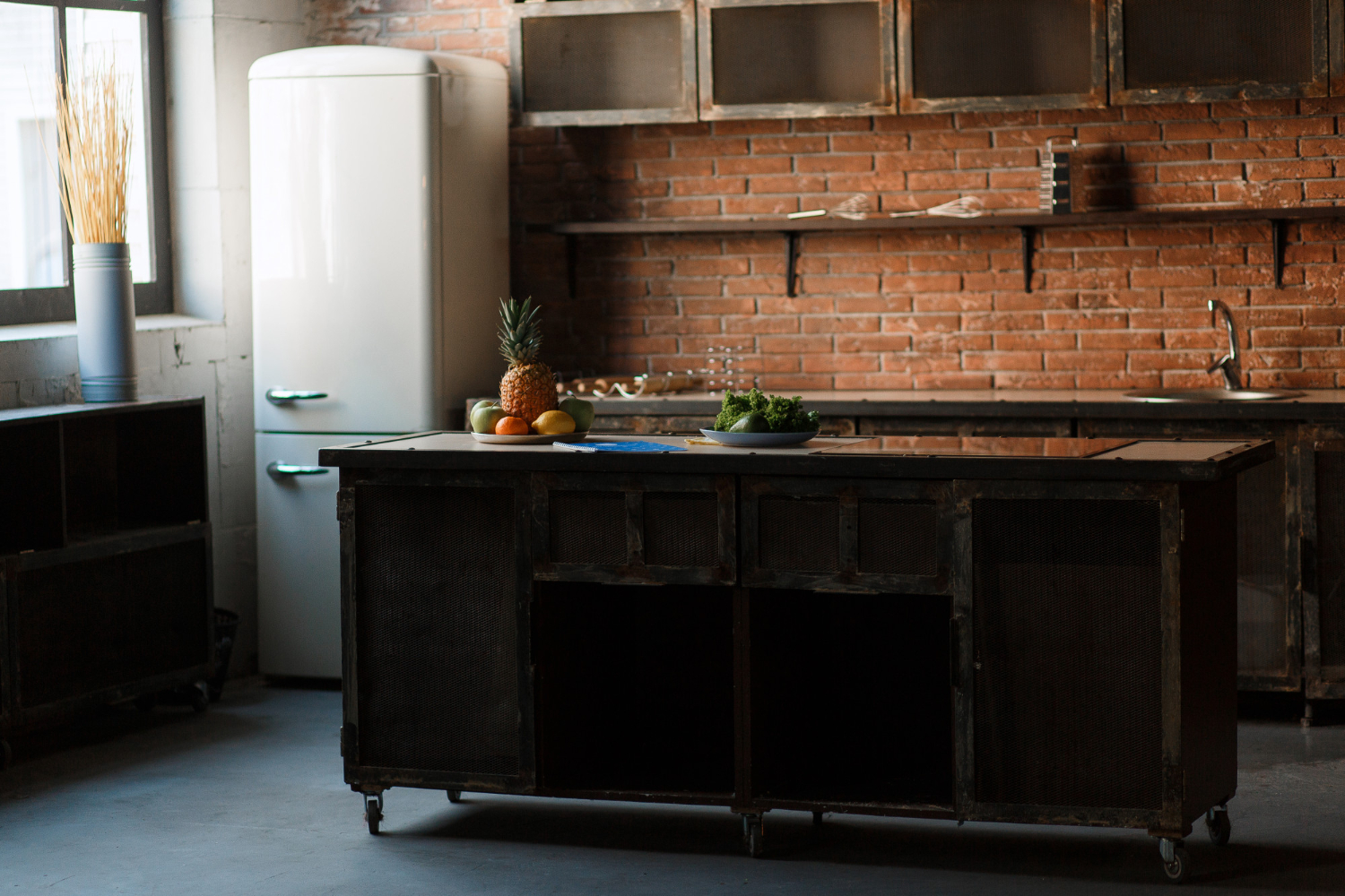 Dated kitchen with exposed red brick walls, dark metal cabinetry, vintage white refrigerator, central island with fruit, and muted natural lighting.