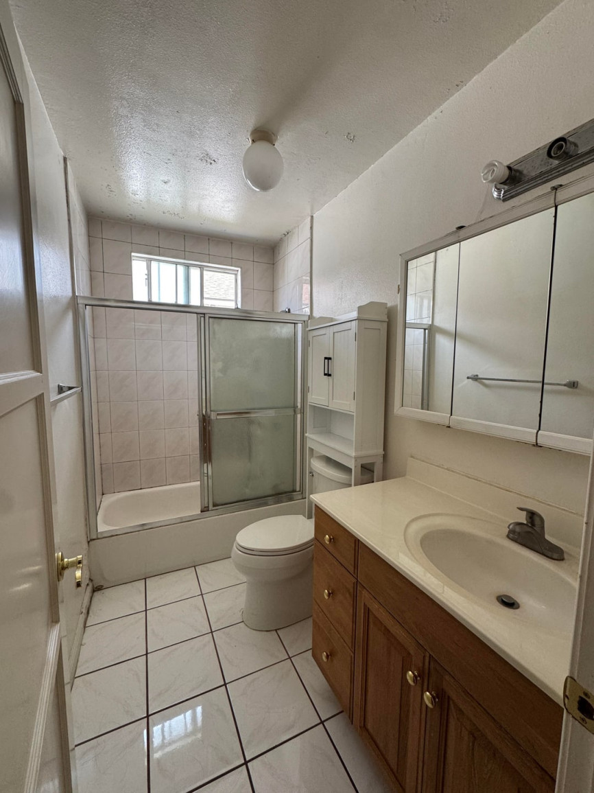Small traditional bathroom with white tiled floor and walls, wooden vanity with cream countertop, frosted glass shower doors, and minimal natural light.