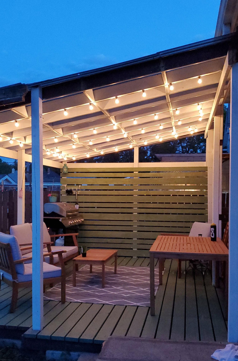 Outdoor patio with wooden deck, beige cushioned chairs, string lights on white pergola, a grill, wooden table, and soft evening ambiance.
