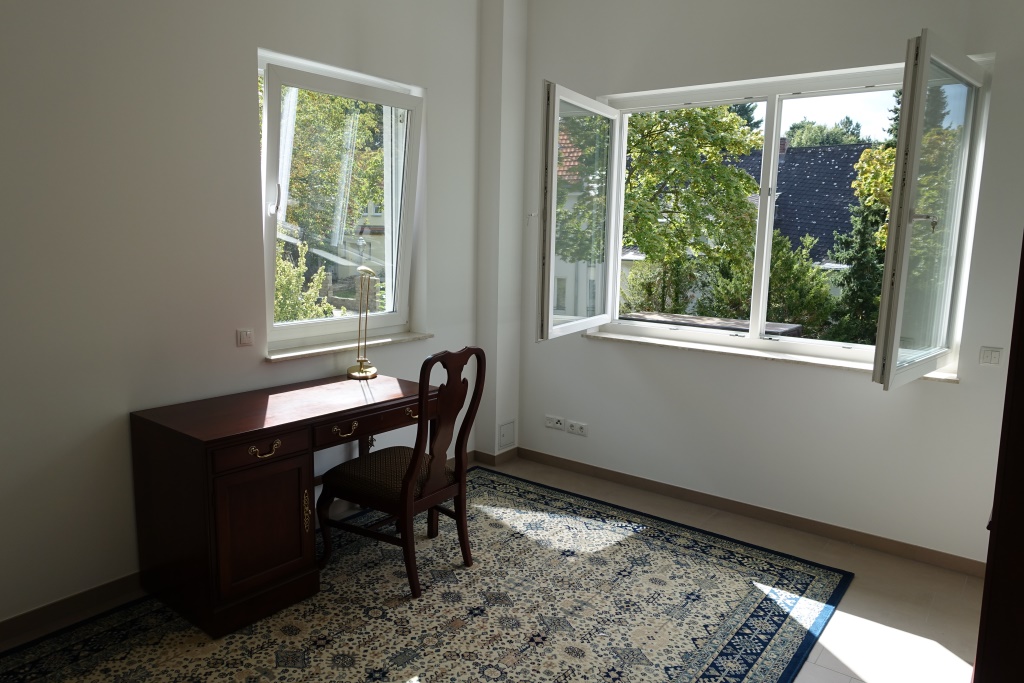 Minimal room with wooden desk, patterned rug, white walls, and large open windows with natural daylight