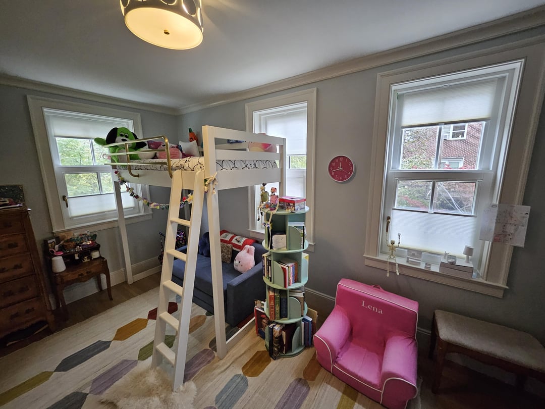 Kids bedroom with white loft bed, pink chair, gray walls, and colorful patterned rug beside large windows
