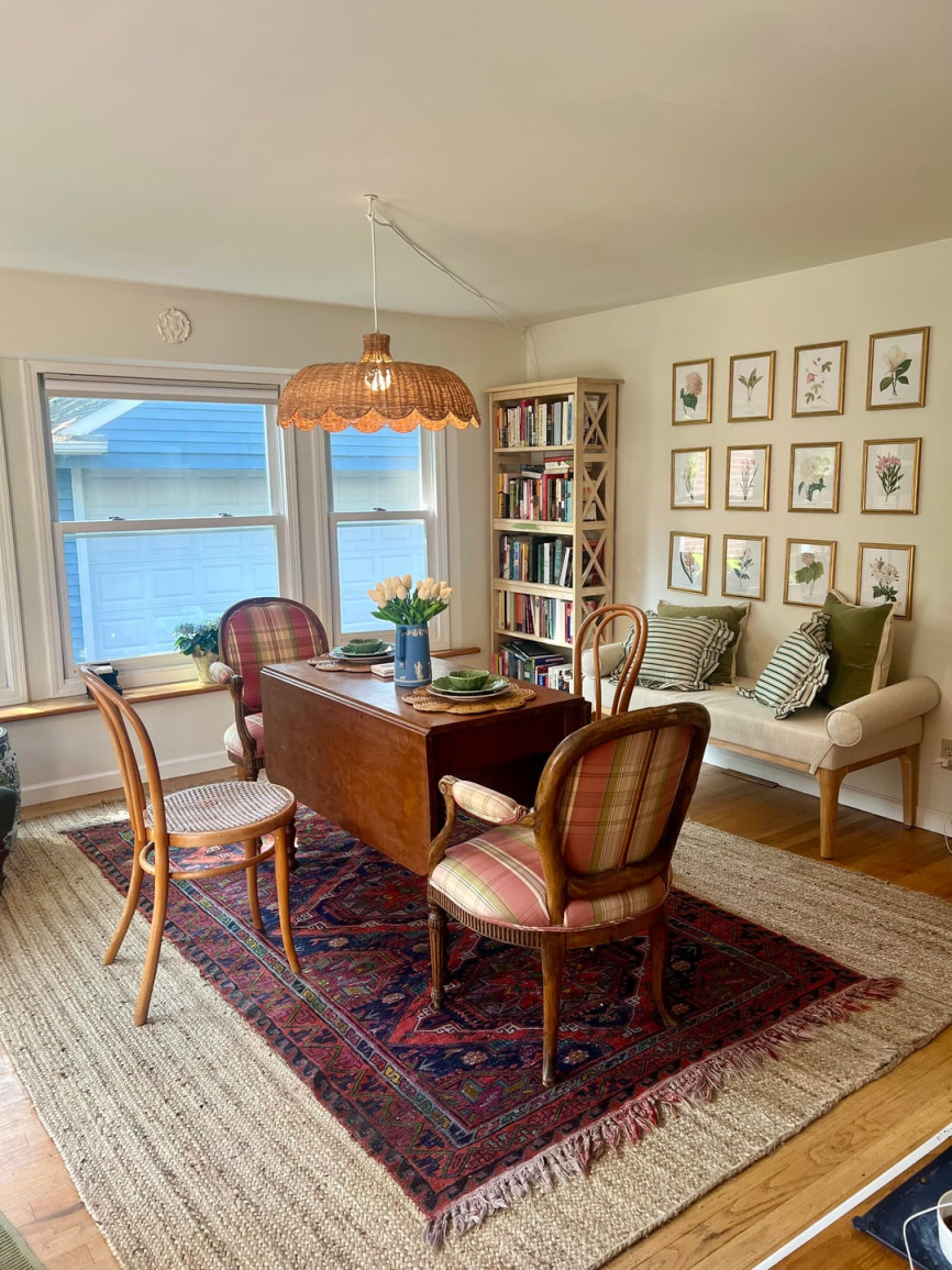 Traditional dining room with wooden table, mixed vintage chairs, layered rugs, botanical wall art, natural light, and a woven pendant light.